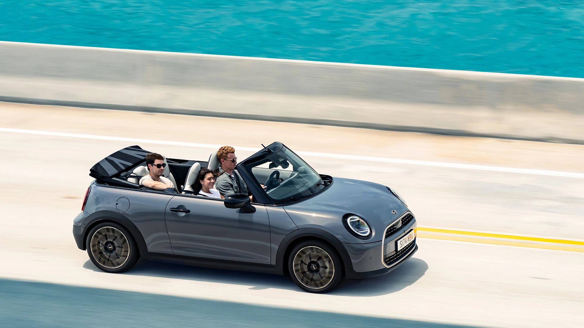 Ein Mini Cooper Cabrio fährt auf einer Straße die an einem Meer angrenzt. Das Verdeck des Fahrzeugs ist geöffnet. Es sitzen drei Personen drinnen. 2 vorne, eine hinten.